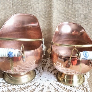 May include: Two copper coal scuttles with brass handles and porcelain knobs. The scuttles have a rounded, oval shape and are sitting on a white lace doily.