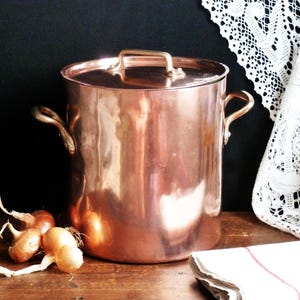 May include: A large, shiny copper pot with a lid and two side handles. The pot is sitting on a wooden surface with a few onions and a white lace cloth in the background. The pot is a beautiful, vintage kitchen item.