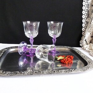 Puede incluir: Tres copas de vino de cristal transparente con tallos morados descansan sobre una bandeja plateada. Una copa está tumbada de lado. Un pequeño racimo de bayas rojas y hojas verdes está en la bandeja.