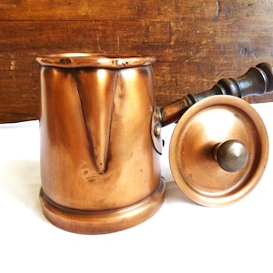May include: A vintage copper pot with a wooden handle and lid. The pot has a rounded base and a cylindrical body with a spout. The lid is round with a brass knob. The handle is dark wood. The pot is sitting on a white surface.
