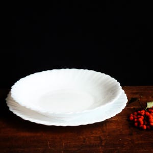 Può includere: Due piatti bianchi con bordo smerlato sono impilati su una superficie di legno scuro. Un grappolo di bacche rosso vivo e una foglia verde si trovano a destra dei piatti. Lo sfondo è nero.
