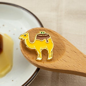 May include: A gold-colored enamel pin shaped like a camel with a brown pudding dessert on its back. The pudding has a green leaf on top and a brown chocolate sauce dripping down the sides.