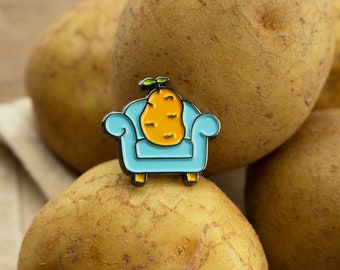 May include: A blue enamel pin featuring a cartoon potato sitting in a blue armchair. The potato is yellow with a green sprout on top. The pin is sitting on top of a pile of potatoes.