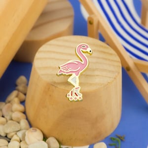 May include: A pink flamingo pin with gold accents, wearing roller skates. The pin is on a wooden surface.