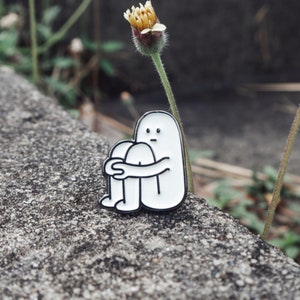 Könnte beinhalten: Weißer Emaille-Pin mit einem Cartoon-Charakter mit traurigem Gesichtsausdruck, der mit seinen Armen um seine Beine gewickelt sitzt. Der Pin befindet sich auf einer grauen, strukturierten Oberfläche.