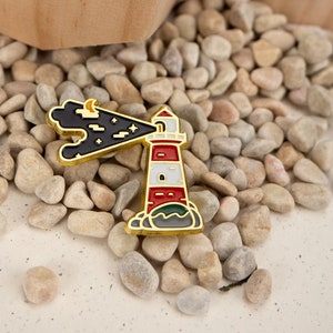 May include: Gold-tone enamel pin depicting a lighthouse with red and white stripes, a black cloud with stars, and a crescent moon. The lighthouse is set against a green background.
