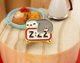 May include: Gold enamel pin featuring a gray sloth sleeping on a white alarm clock with the text "Z:ZZ".