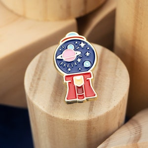 Könnte beinhalten: Eine goldfarbene Emaille-Anstecknadel in Form einer Gummibärchen-Maschine mit blau-weißem Design. Die Gummibärchen-Maschine ist mit Sternen und Planeten gefüllt.