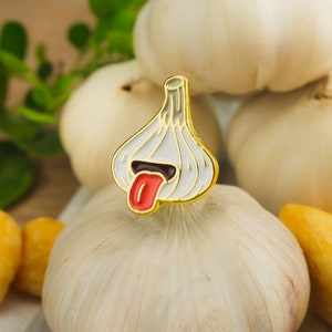 May include: A gold-tone enamel pin shaped like a garlic clove with a red tongue sticking out. The pin is sitting on top of a white garlic bulb.
