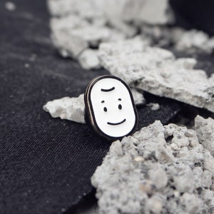 May include: A white enamel pin with a black outline of a smiling face. The pin is on a grey and black background.