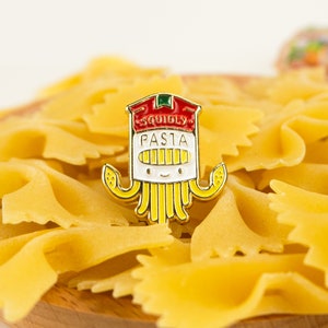 May include: A gold-colored enamel pin shaped like a cartoon octopus wearing a chef's hat. The hat says "Squidly Pasta". The pin is sitting on a bed of uncooked pasta.