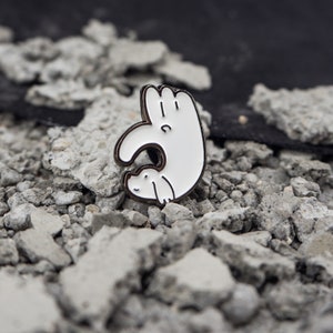 May include: White enamel pin of a hand making an okay gesture with a small white dog inside the hand.
