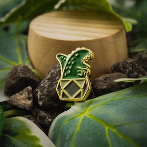 May include: A gold-colored enamel pin featuring a green dinosaur in a geometric pot. The dinosaur is depicted with a toothy grin and a green, scaly body.