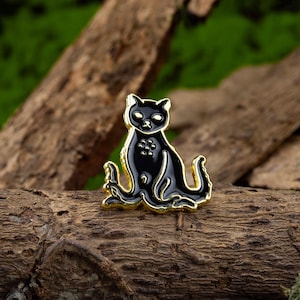 May include: Black enamel pin of a cat with a curled tail, sitting on a wooden surface. The cat has a gold outline and black eyes.