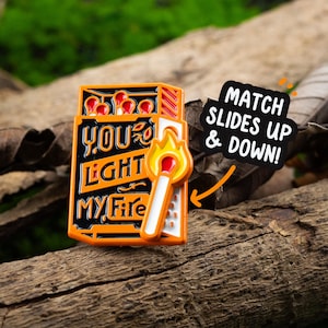 May include: Orange and black enamel pin with a matchbox design. The matchbox says "You Light My Fire" and has a matchstick that slides up and down. The text "Match Slides Up & Down!" is in a speech bubble.