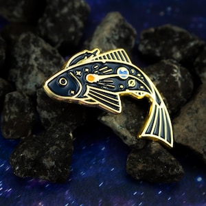 May include: A gold-toned enamel pin shaped like a fish with black and gold details. The fish has a celestial design with planets and stars on its body.
