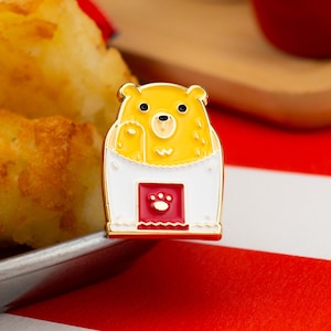 May include: A yellow bear enamel pin with a white and red border. The bear is waving from a white container with a red paw print at the bottom. The pin has a gold outline and black eyes.