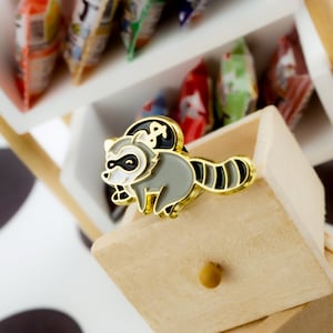 May include: A gold-colored enamel pin featuring a cartoon raccoon with black eyes, a white face, and a gray body. The raccoon is wearing a black and white striped tail.