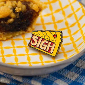 May include: A yellow and red enamel pin shaped like a pie slice with the word "SIGH" written on it. The pin is sitting on a white and yellow patterned plate.