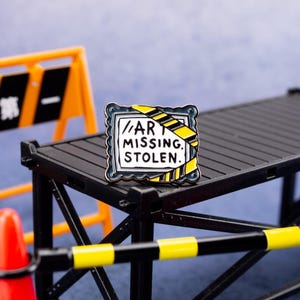 May include: A rectangular enamel pin with a black border and the words "ART MISSING. STOLEN." in a white box. The pin has a yellow and black striped design on the right side, resembling caution tape. The pin is set against a black structure with an orange and yellow barrier in the background.