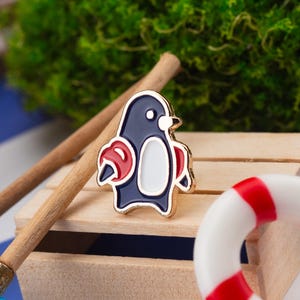 May include: A navy blue and white penguin enamel pin with red accents and a gold border. The pin is set against a wooden background with a miniature life preserver and a wooden stick.