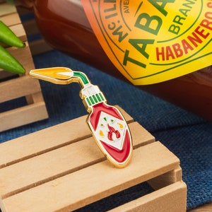 May include: Gold-colored enamel pin shaped like a hot sauce bottle with a red and white label. The label has a flame design and the text "TABASCO BRAND HABANERO" is visible.