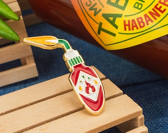 May include: Gold-coloured enamel pin shaped like a hot sauce bottle with a red and white label. The label has a flame design and the text "TABASCO BRAND HABANERO" is visible.