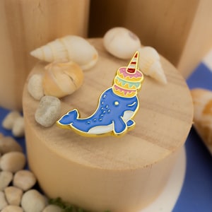 May include: A gold-tone enamel pin of a blue narwhal with a white belly and a pink, green, and white donut on its head.