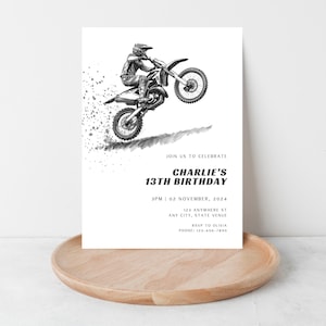 May include: A black and white birthday invitation featuring a motocross rider on a motorcycle. The invitation includes the text "CHARLIE'S 13TH BIRTHDAY" with event details. The invitation is displayed on a wooden tray.