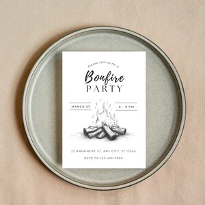 May include: A white invitation card with the words "Bonfire PARTY" in a stylish font, featuring a bonfire illustration. The card includes the date, time, and RSVP information. The invitation is placed on a gray plate.