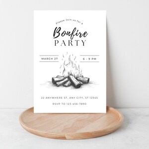 May include: A white invitation card with the words "Bonfire PARTY" in a stylish font, along with the date and time. A detailed illustration of a bonfire is featured. The card is placed on a wooden tray.