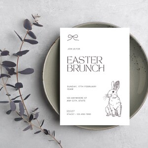 May include: An Easter brunch invitation on a white card with a bunny illustration, set on a green plate. The card reads "EASTER BRUNCH" with event details. A sprig of dark green leaves is on the left.