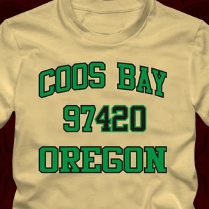 Könnte beinhalten: Ein beiges T-Shirt mit grünem Text, der "COOS BAY 97420 OREGON" lautet, und den Worten "Yellow Haze" in gelber Schrift unten.