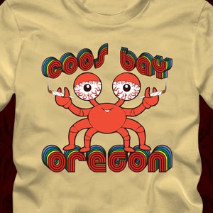 Puede incluir: Una camiseta beige con un gráfico de un cangrejo rojo con ojos grandes que sostiene dos cigarrillos. El cangrejo está encima del texto "Coos Bay Oregon" en colores arcoíris. El texto "Yellow Haze" está en la parte inferior de la imagen.