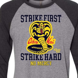 May include: Grey and black baseball tee shirt with a yellow cobra logo and the text "Strike First Strike Hard No Mercy" and "Cobra Kai The Karate Kid".