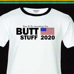 May include: White t-shirt with a black ringer neck. The shirt has a black graphic that says "Time To Try Something New" and "BUTT STUFF 2020" with an American flag.