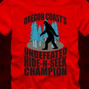Puede incluir: Camiseta roja con una silueta negra de Bigfoot en un bosque, el texto "Oregon Coast's Undefeated Hide-N-Seek Champion" está impreso en la camiseta.