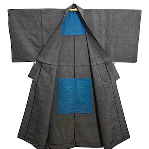 Puede incluir: Un kimono gris con interior y detalles azules, colgado de una percha de madera. El kimono está abierto, revelando la tela azul. Las mangas son anchas y la tela parece texturizada.