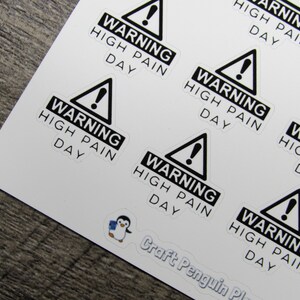 May include: A sheet of white paper with multiple black and white warning stickers. The stickers feature a black triangle with an exclamation point and the text "WARNING HIGH PAIN DAY".