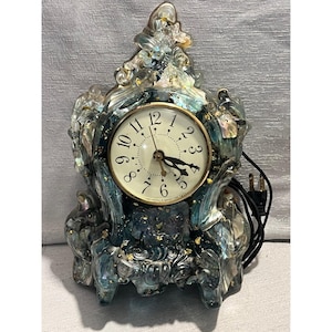 May include: A decorative electric mantel clock featuring a translucent, swirling blue and iridescent resin frame embedded with gold leaf flakes. The clock face is round with a gold-tone bezel, black Arabic numerals, and black hands.