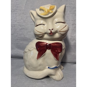 May include: A ceramic cat-shaped cookie jar with a glossy off-white finish. The cat wears a yellow hat with a bird detail, a large dark red bow at the neck, and has painted brown whiskers and closed eyes. A thin blue line accents the base.