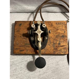 Op de afbeelding: Een vintage telegraafsleutel gemonteerd op een rechthoekige houten basis met zichtbare markeringen en krassen. Het apparaat heeft een zwart metalen frame, een zilverkleurige metalen hendel en een ronde zwarte knop. Een bruin snoer is aan de bovenkant bevestigd.