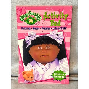May include: A Cabbage Patch Kids Activity Pad featuring a doll with dark skin, black hair in pigtails with light purple bows, and a striped shirt. The pink cover displays the text Cabbage Patch Kids Activity Pad, Coloring, Mazes, Puzzles, Lots of FUN!, and Stickers Included, along with a small brown teddy bear.