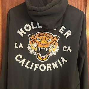 May include: A black hooded jacket featuring an embroidered graphic on the back. The design includes a snarling leopard head in orange, black, and white, surrounded by the white embroidered text HOLLISTER LA CA CALIFORNIA in a circular layout.