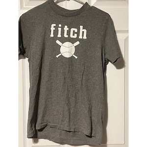 May include: A heather grey short-sleeve t-shirt featuring a crew neckline and a white graphic print on the chest. The graphic displays the word fitch in lowercase letters above a white baseball icon crossed by two white baseball bats.