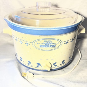 May include: A Rival Crock-Pot stoneware slow cooker with a cream-colored base featuring blue speckled accents and a blue floral pattern. It includes a clear glass lid, two side handles, a front control knob for off, low, and high settings.
