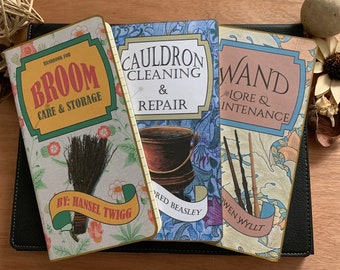 May include: Three hardcover books with titles "Broom Care & Storage", "Cauldron Cleaning & Repair", and "Wand Lore & Intenance". The books have floral and botanical designs with text in a vintage style. The books are on a black surface.