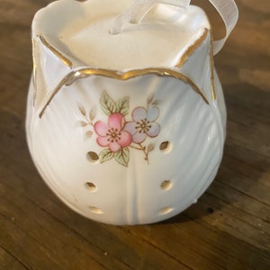 May include: A white ceramic ornament with a floral design in pink and light blue. The ornament has a scalloped edge with gold trim and a sheer ribbon for hanging. Small holes are evenly spaced around the bottom.
