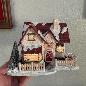 May include: A miniature Christmas-themed cottage with a red roof and snow-covered details. The cottage has lit windows, a small fence, and a sign that reads "Candy Cane Cottage". A small snowman figure is also present.