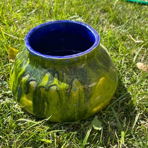 May include: A round ceramic vase with a wide opening and a bulbous body. The exterior is a blend of green and yellow with dark green streaks, while the interior is a deep blue. The vase is sitting on a bed of green grass.